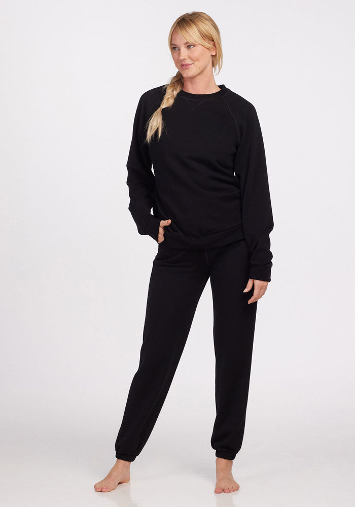 A person with long blonde hair in a side braid stands barefoot against a white background, wearing a black long-sleeve sweatshirt and Woolx Parker Sweatpants in Black for optimal temperature regulation.