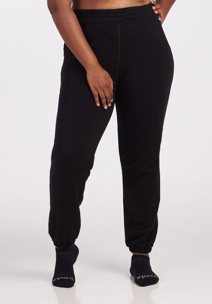 A person stands against a plain background wearing Woolx Parker Sweatpants in black and black socks, with their left hand resting on their right thigh. Only the lower half of their body is shown. | Le'Quita is 5'11", wearing a size XL