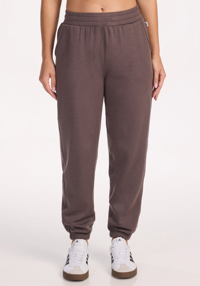 A person wears Woolx Parker Sweatpants in Plum Truffle—brown joggers with an elastic waistband and cuffs—paired with white athletic shoes, standing against a plain white background with hands relaxed at their sides.