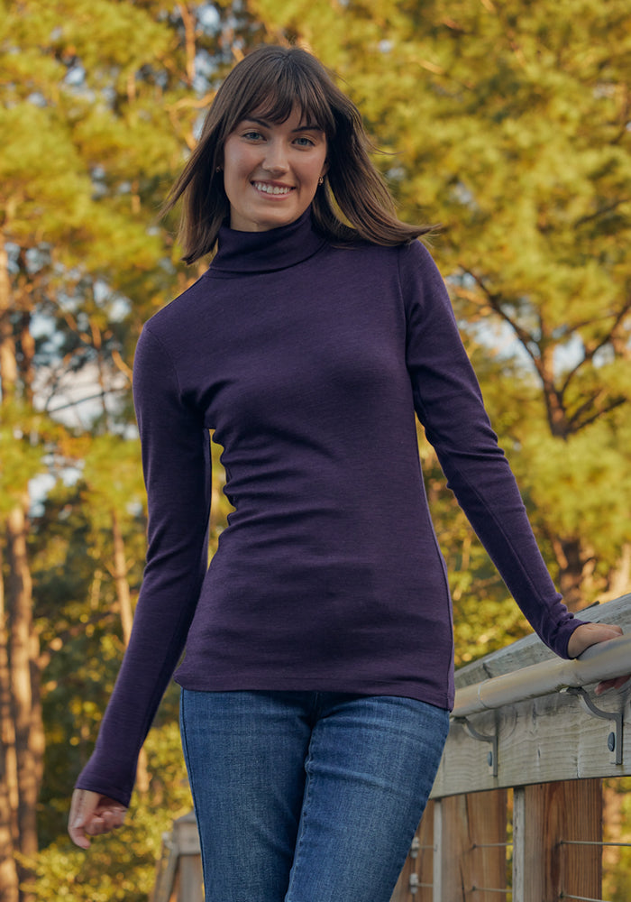 A woman with shoulder-length brown hair, wearing the Woolx Peyton Turtleneck in Deep Plum and jeans, stands smiling on a wooden bridge with green trees behind her.