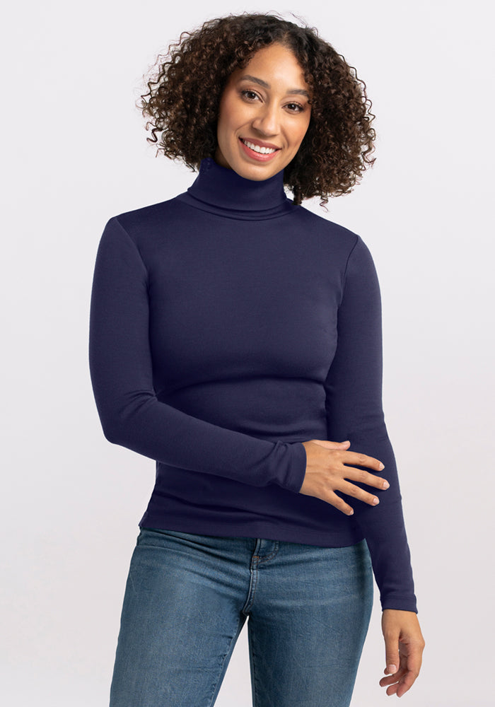 A woman with curly hair smiles, wearing the Woolx Peyton Turtleneck in Midnight Sapphire and blue jeans, standing against a plain light background.