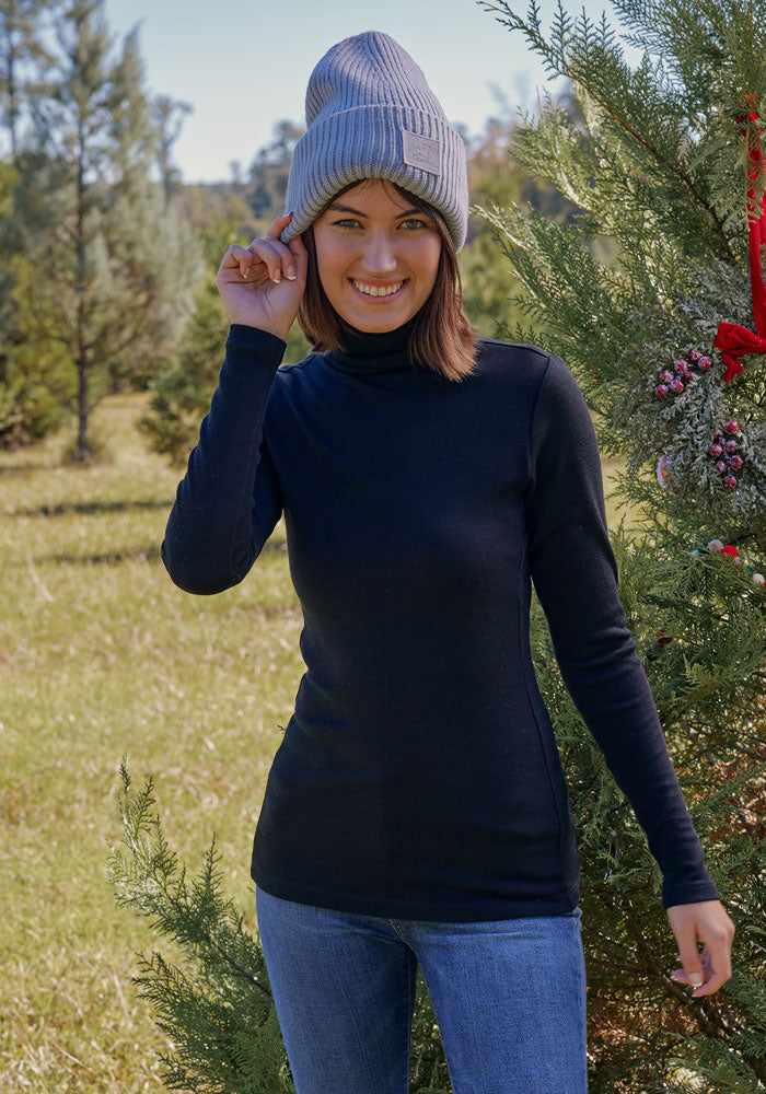 A woman wearing a gray beanie, blue jeans, and the Woolx Peyton Turtleneck in Black smiles while posing outdoors by a decorated pine tree on a sunny day.