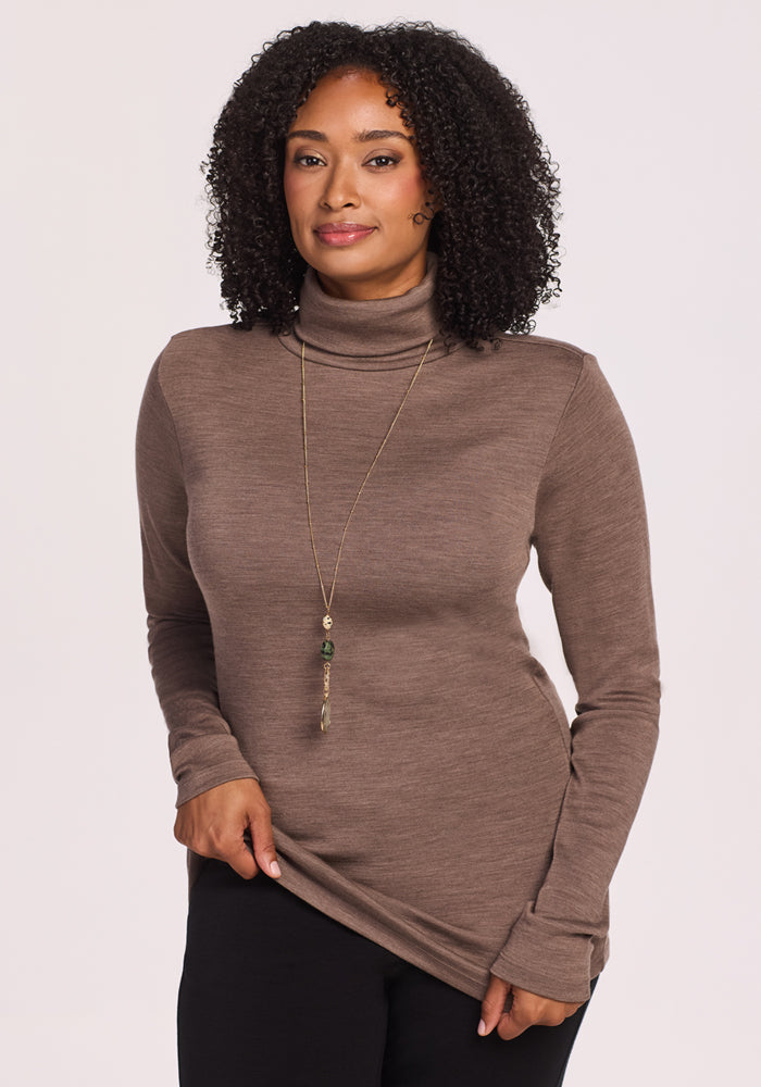 A woman with curly hair wears the Woolx Peyton Turtleneck in Simply Taupe, holding the hem of her sweater and smiling gently at the camera against a plain light background.