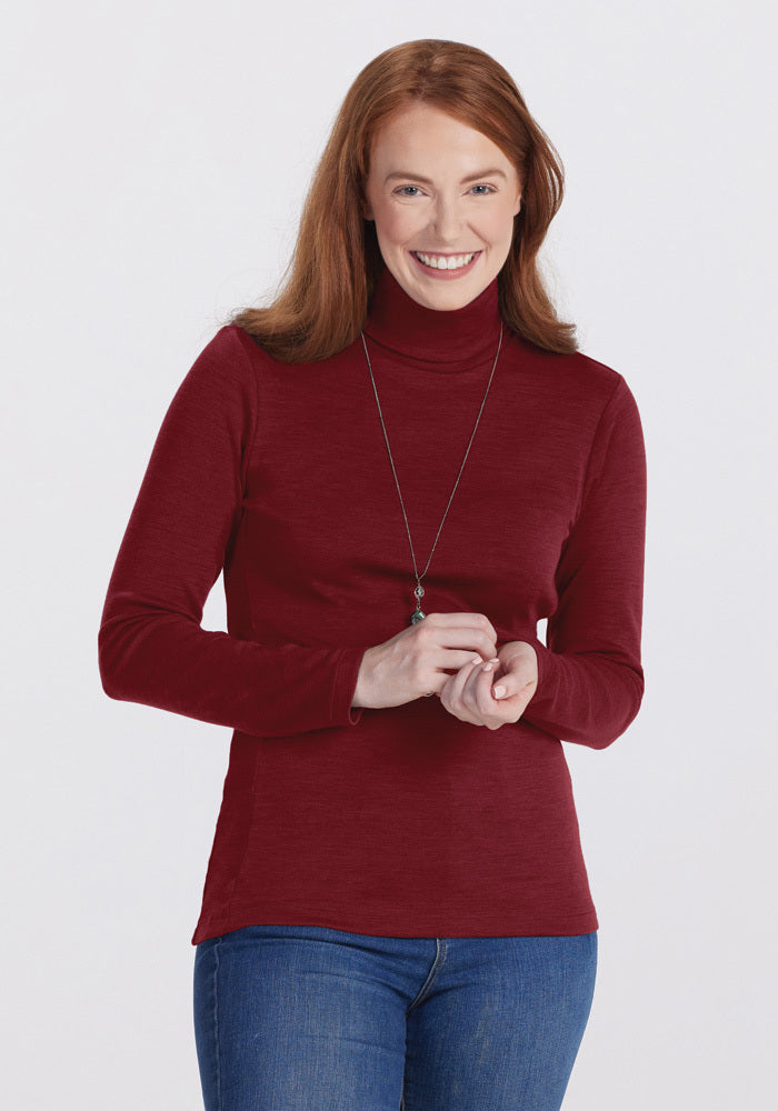A smiling woman with long red hair wears the Woolx Peyton Turtleneck in Sweet Pepper and blue jeans, accessorized with a long necklace. She stands against a plain white background.