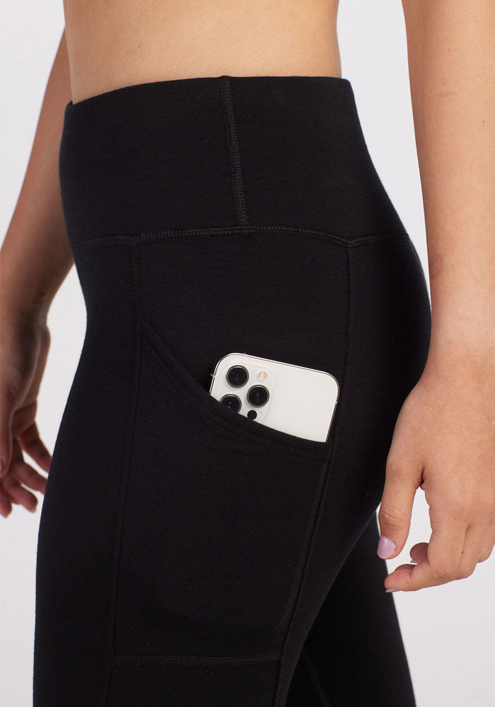 A person wearing the Woolx Piper Pocket Leggings in black, featuring a moisture-wicking fabric, has their smartphone conveniently tucked into the side pocket. The high-waisted design subtly highlights the phone's camera lenses, while their relaxed hand by their side exudes comfort. These leggings are ideal as a cold-weather baselayer for those chilly days.