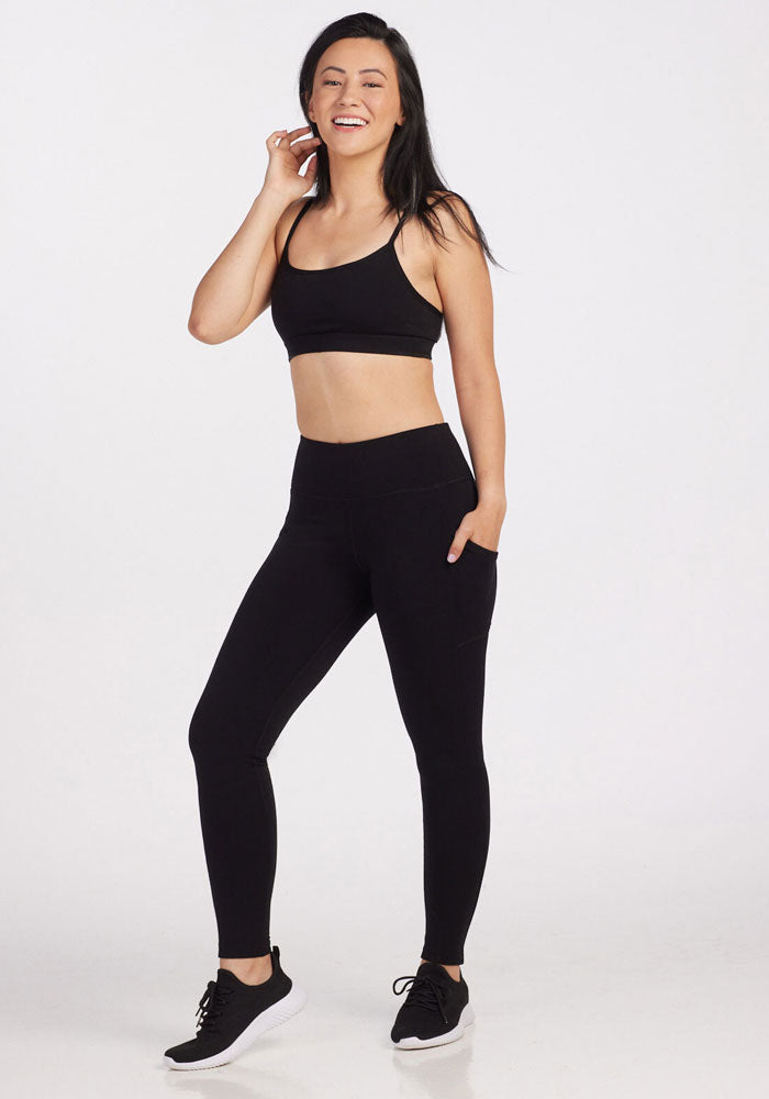 A woman in black athletic wear, featuring a moisture-wicking sports bra and Piper Pocket Leggings by Woolx, smiles while standing against a plain white background. She has one hand in her pocket and is wearing black sneakers.