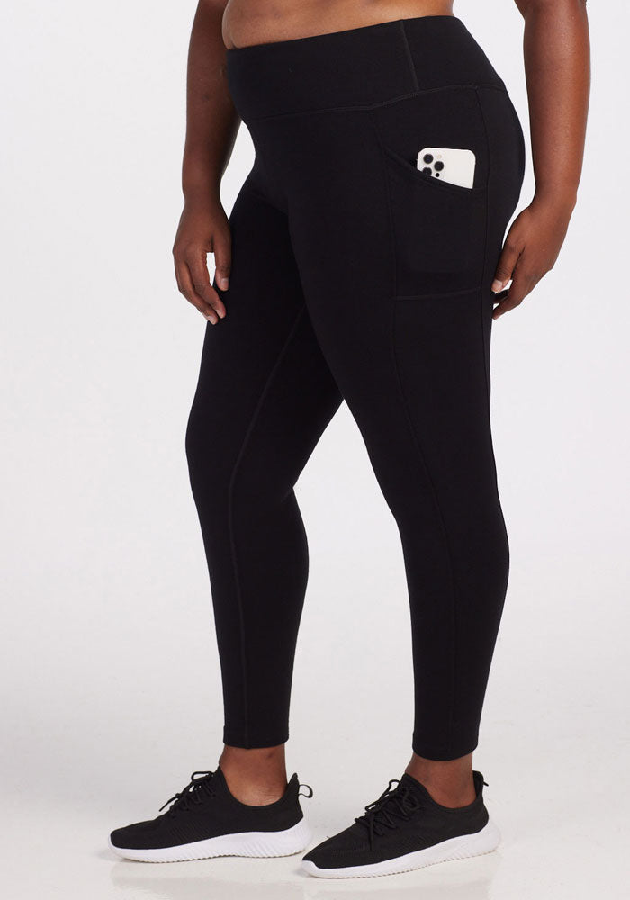 A person is wearing the Woolx Piper Pocket Leggings in black, featuring a convenient side pocket for a smartphone. They are paired with black sneakers, making this ensemble an ideal cold-weather baselayer choice against a plain white backdrop.