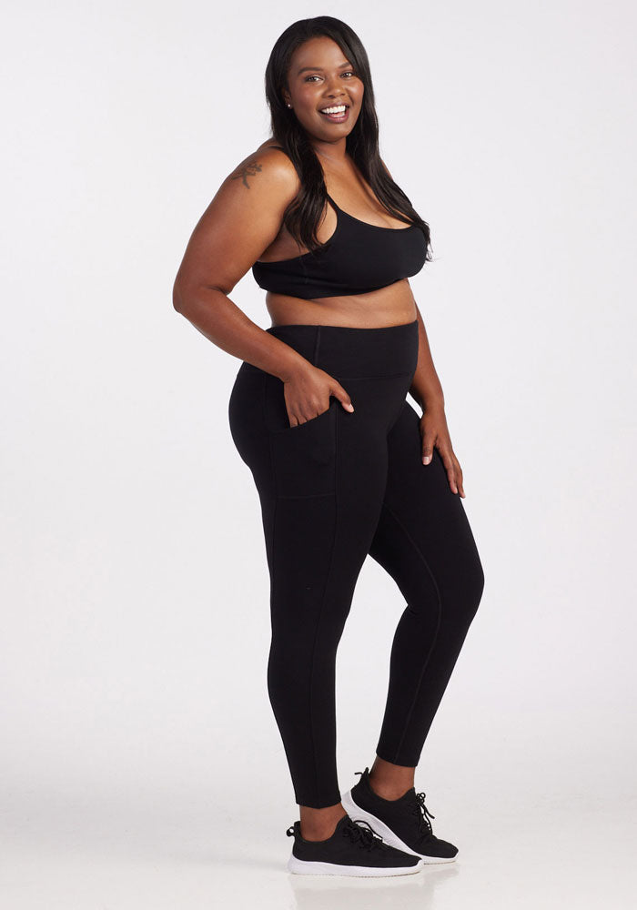 A woman is smiling and posing in her athletic wear, featuring a black moisture-wicking sports bra, Woolx's Piper Pocket Leggings in Black—ideal as a cold-weather baselayer—and sleek black sneakers against a plain white background.