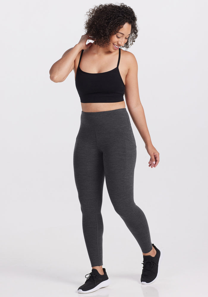 A woman with curly hair smiles while standing on one leg, wearing a black crop top, Woolx Piper Pocket Leggings in Cinder Melange, and black sneakers against a plain white background.