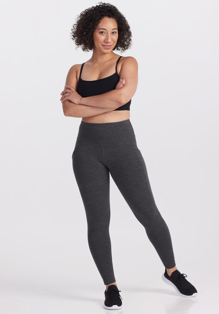 A woman with curly hair stands confidently with arms crossed, wearing a black sports bra, black sneakers, and Woolx Piper Pocket Leggings in Cinder Melange against a plain white background.