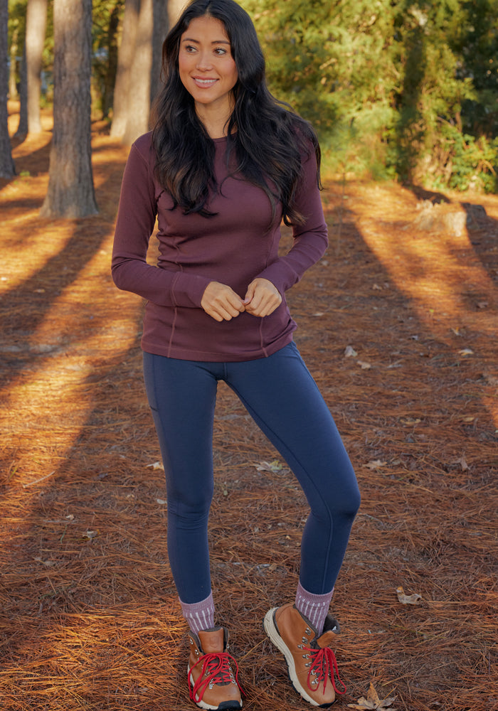 A woman stands outdoors on a pine-covered forest floor, wearing Woolx Piper Pocket Leggings in Deep Navy and a maroon long-sleeve top. Patterned socks and brown hiking boots with red laces complete her look amid sunlit trees.
