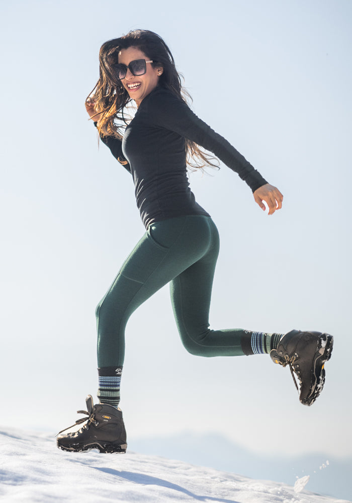A woman in sunglasses, a black long-sleeve top, Woolx Piper Pocket Leggings in Deep Timber, striped socks, and hiking boots jumps playfully on a snowy mountain under a bright sky.