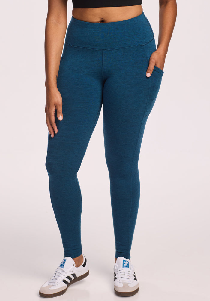 A woman stands against a plain light background wearing Woolx Piper Pocket Leggings in Majolica Blue, a black crop top, and white sneakers with black stripes—an ideal women’s baselayer look for comfort and style.