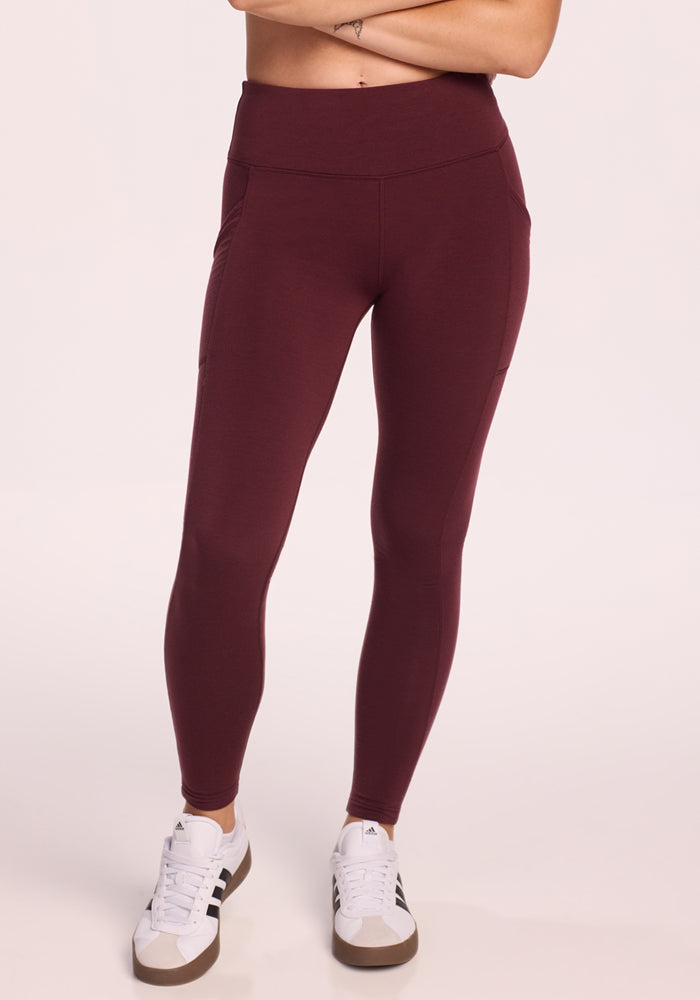 A person stands against a light background in Woolx Piper Pocket Leggings • Final Sale - Toasted Garnet paired with white sneakers. These high-waisted leggings feature side pockets and provide women with comfort and style. Arms are cropped out of frame.