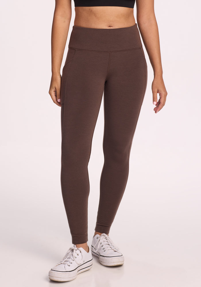 A woman stands against a plain light background wearing Woolx Piper Pocket Petite leggings in Bracken, paired with a black crop top and white sneakers. Only her body from shoulders down is visible.