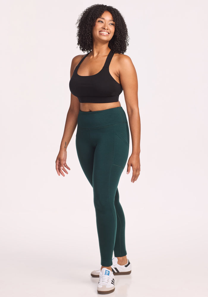 A woman with curly hair smiles in the Woolx Piper Pocket Petite leggings in Deep Timber, styled for cold weather. She wears a black sports bra and white sneakers, standing relaxed against a plain light background.