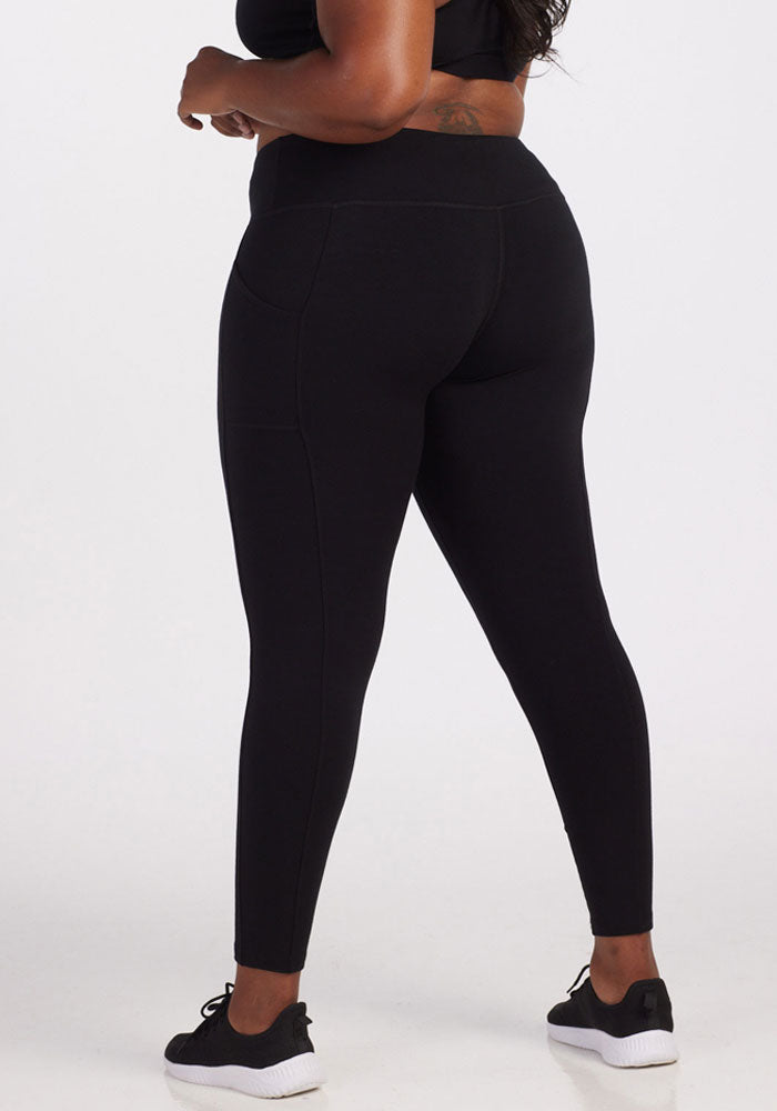 A person stands barefoot against a plain white background, wearing snug Piper Pocket Tall - Black leggings and a matching top from Woolx. Designed for tall women, the leggings offer a sleek silhouette with their elevated waistline.