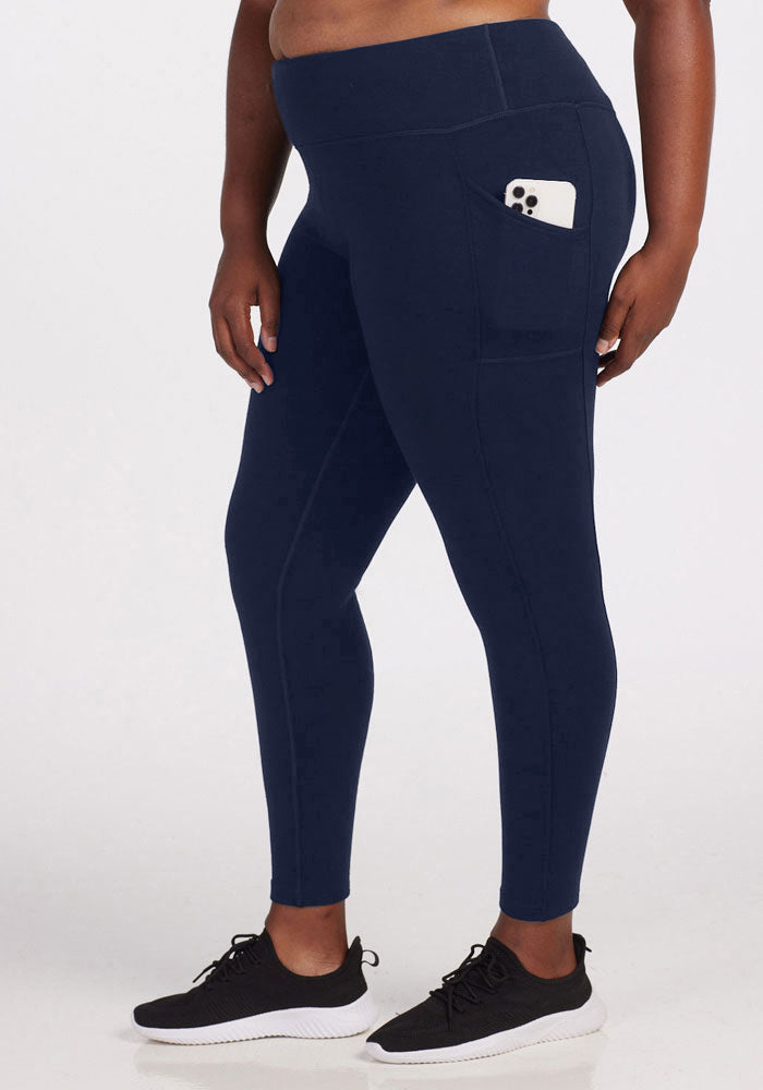 A person stands against a plain white background wearing Woolx Piper Pocket Tall leggings in Deep Navy, featuring a side pocket holding a smartphone, paired with black sneakers.