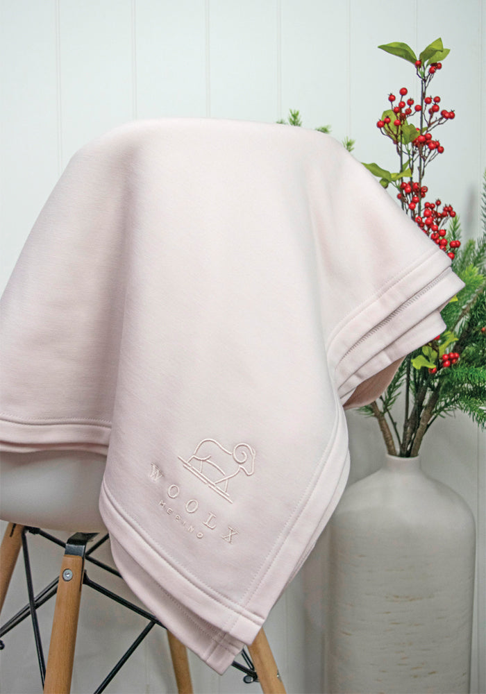 The Placid Merino Blanket - Almost Mauve by Woolx, featuring an embroidered sheep logo and “Wooly,” is draped over a wooden chair. A white vase with festive greenery and red berries in the background completes this cozy scene.