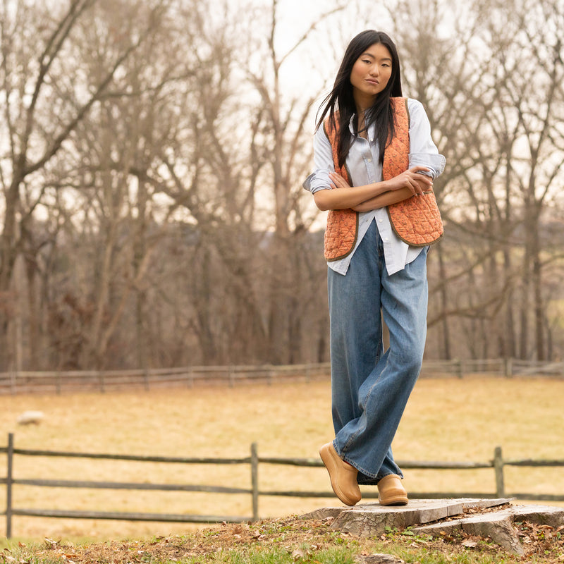 Woman standing outdoors wearing a bright vest, blue button down, jeans, and honey platform Dansko clogs.
