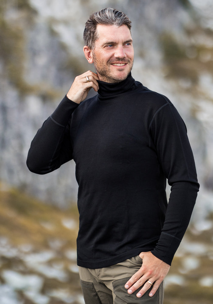 A man with short, dark hair and gray streaks smiles outdoors in tan pants and the Woolx Prescott Turtleneck in black. He touches his collar while standing before a blurred rocky, green background.
