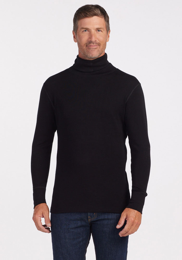 A person with short hair and light stubble is wearing a Woolx Prescott Turtleneck in black, made from soft Merino wool, along with blue jeans. They are standing against a plain white background and looking at the camera with a slight smile. | Lee is 6', wearing a size L
