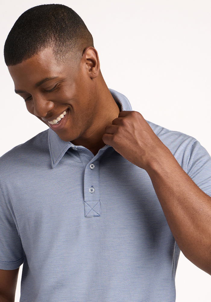 A man wearing a Woolx Preston Polo - Blue Sand Mini Stripe, short-sleeve, looks down and smiles while holding the collar with one hand against a plain light background.