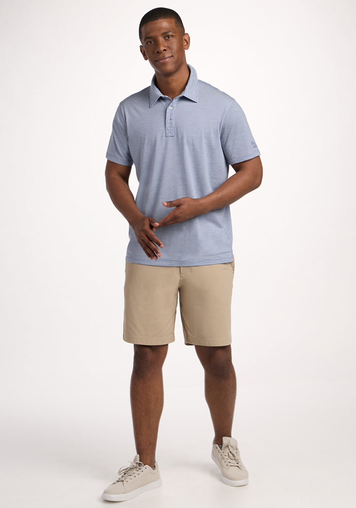 A man stands against a plain white background wearing the Woolx Preston Polo in Blue Sand Mini Stripe, beige shorts, and light sneakers. He faces forward with a slight smile, both hands held in front of him.