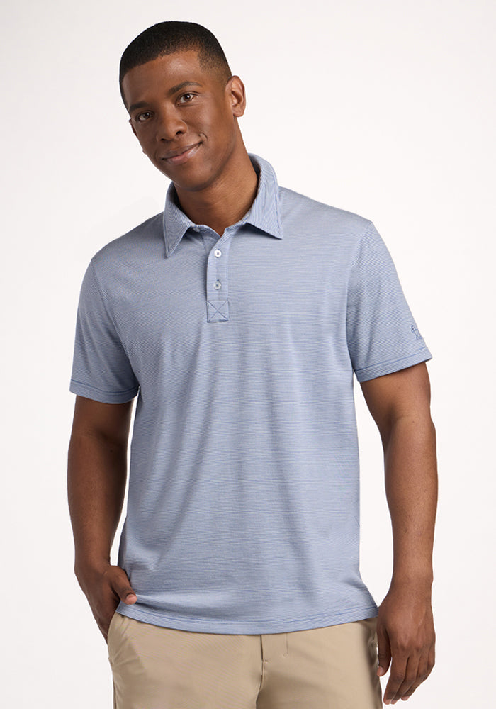 A man stands against a plain background, wearing the Woolx Preston Polo in Blue Sand Mini Stripe—a moisture-wicking shirt—and beige pants, with his hands by his sides and a slight smile on his face.