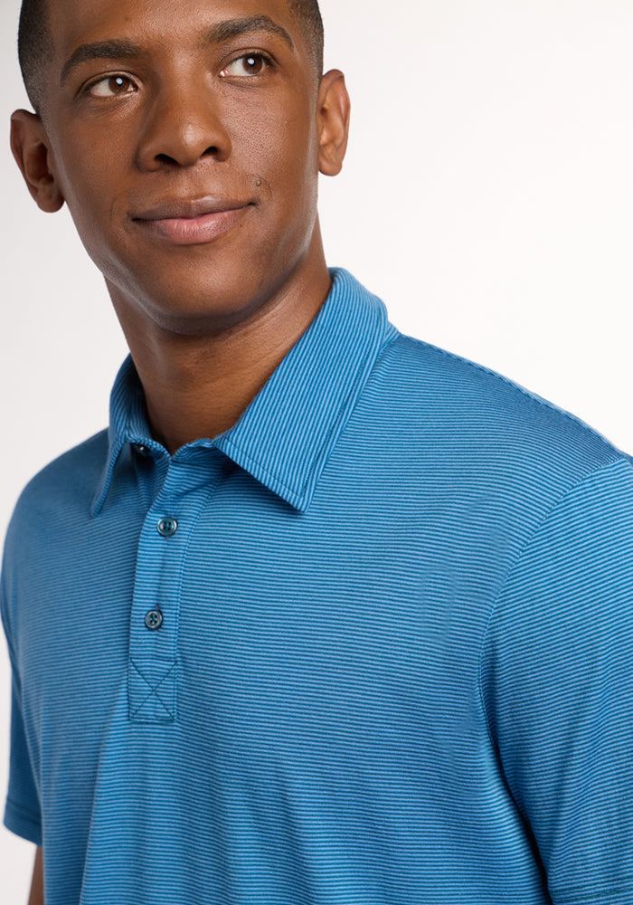 A man in a blue Preston Polo - Coastal Mini Stripe by Woolx, with short sleeves and buttons, stands against a plain light background, looking slightly left and smiling softly.