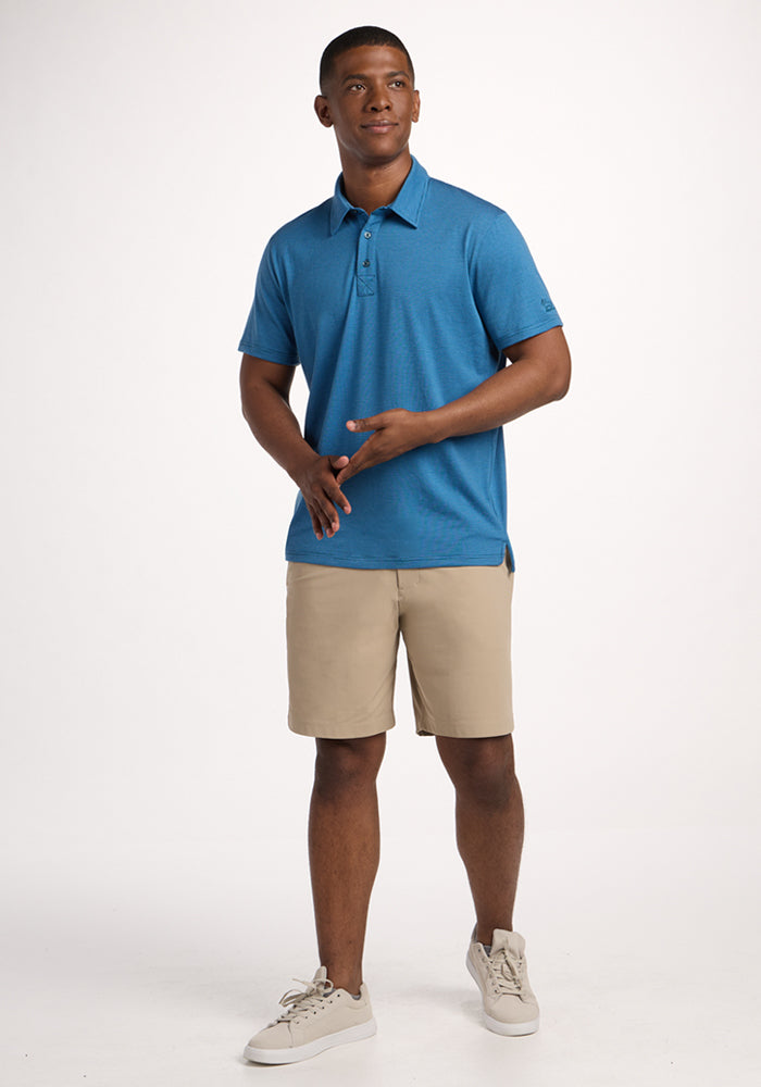 A man stands against a white background, smiling in the Woolx Preston Polo - Coastal Mini Stripe, paired with beige shorts and light sneakers. His relaxed pose highlights the comfort of this moisture-wicking shirt.