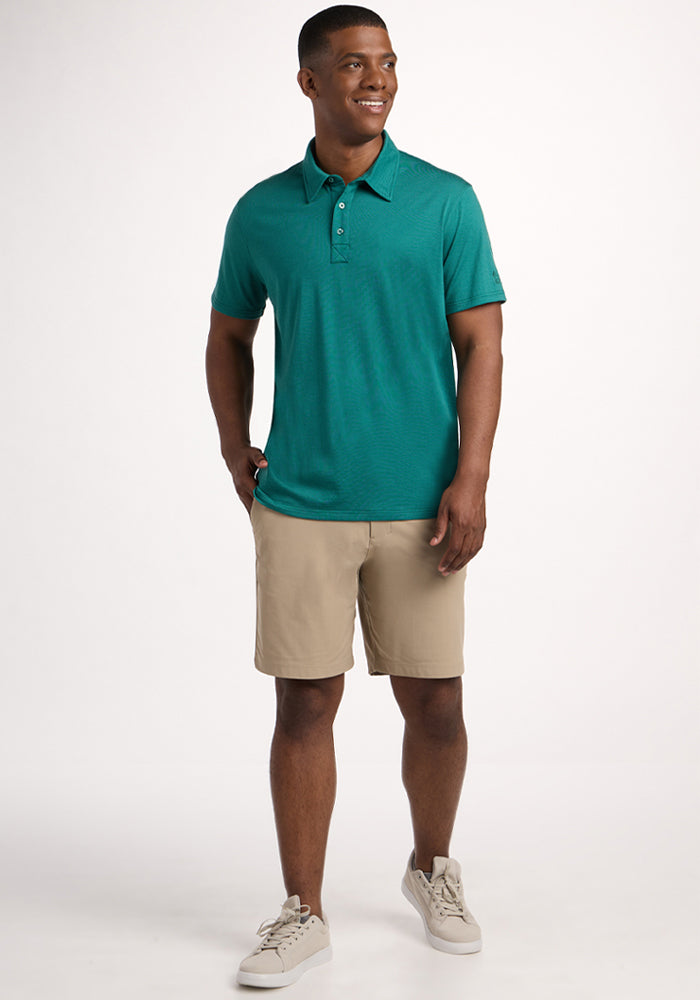 A man smiles in the Woolx Preston Polo - Woodland Mini Stripe, beige shorts, and light sneakers, standing against a white background with one hand in his pocket as he looks to the side.