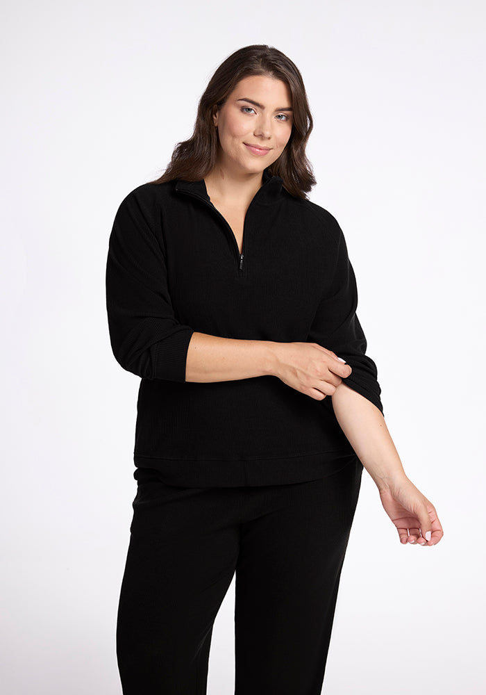 A person with long brown hair is smiling gently while wearing the Woolx Quinn Ribbed 1/4 Zip in black along with black pants. They are rolling up their left sleeve against a plain white background. | Juliana is 5'10", wearing a size XL