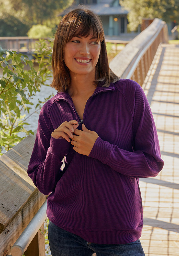A woman with shoulder-length brown hair smiles on a wooden bridge, wearing the Woolx Quinn Ribbed 1/4 Zip in Majestic Purple. Trees and a building appear in the background.