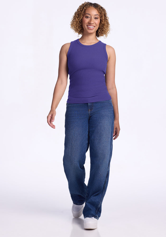 Smiling woman with curly hair wears the Woolx Raegan Ribbed Tank Top in Berry Indigo, blue jeans, and white shoes.