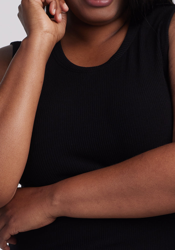 A person wearing a Woolx Raegan Ribbed Tank Top - Black is shown from the shoulders down. One arm crosses the chest while the other hand gently touches the cheek against a plain white background.
