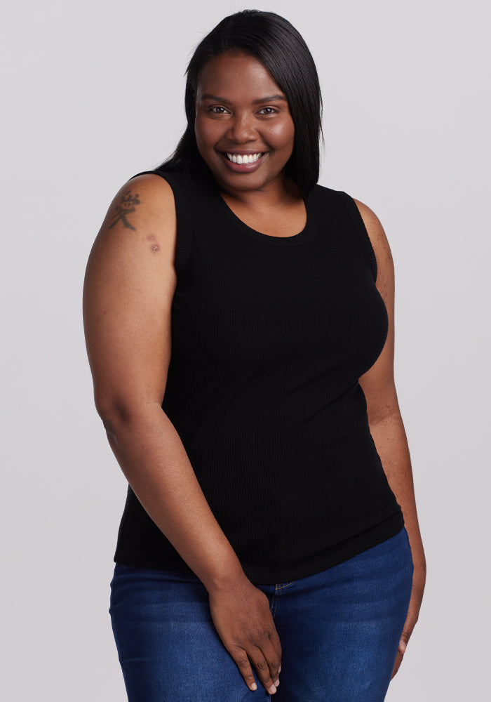 A person with long black hair smiles warmly, dressed in a sleek Woolx Raegan Ribbed Tank Top - Black and blue jeans. They have a tattoo on their left upper arm against a plain gray background. | Le’Quita is 5’11”, wearing a size XL