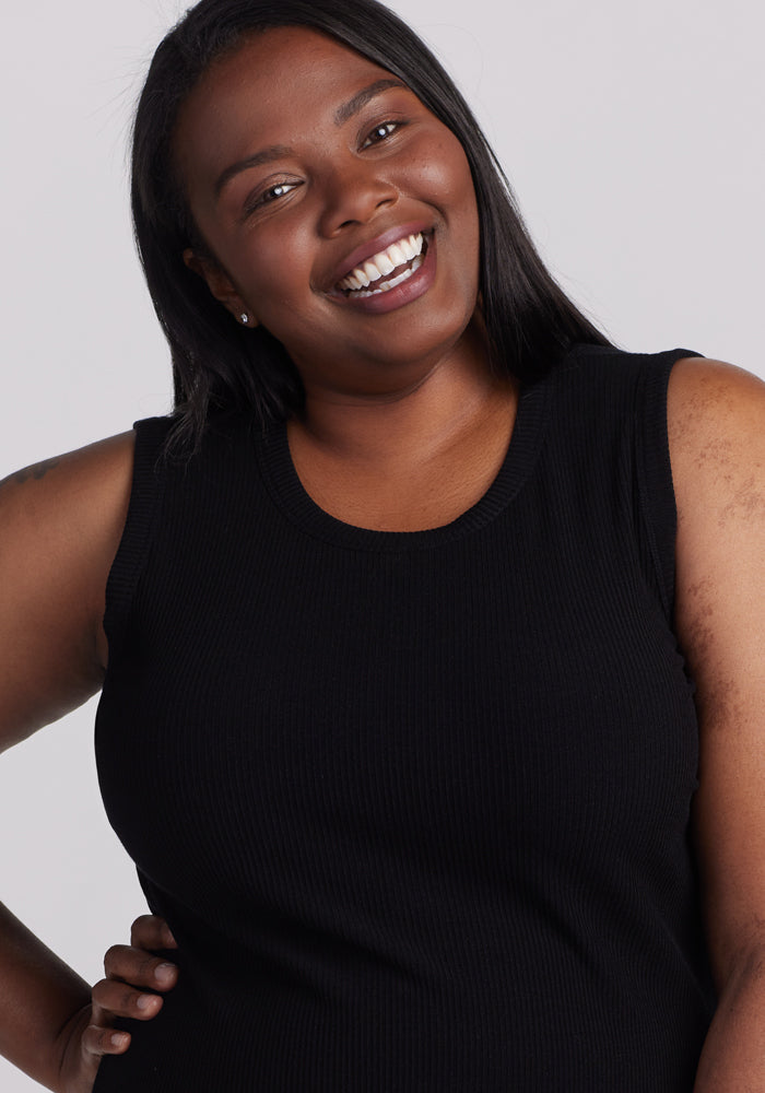 A woman with long hair smiles brightly at the camera, wearing a Woolx Raegan Ribbed Tank Top in black. She has one hand on her hip, exuding confidence against a plain gray backdrop.