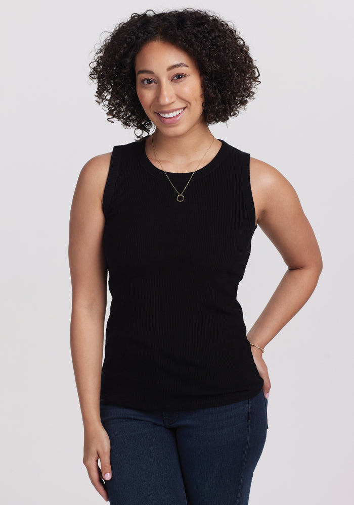 A person with curly hair wearing a Woolx Raegan Ribbed Tank Top in black and blue jeans smiles while standing against a plain background. | Tori is 5'7", wearing a size S