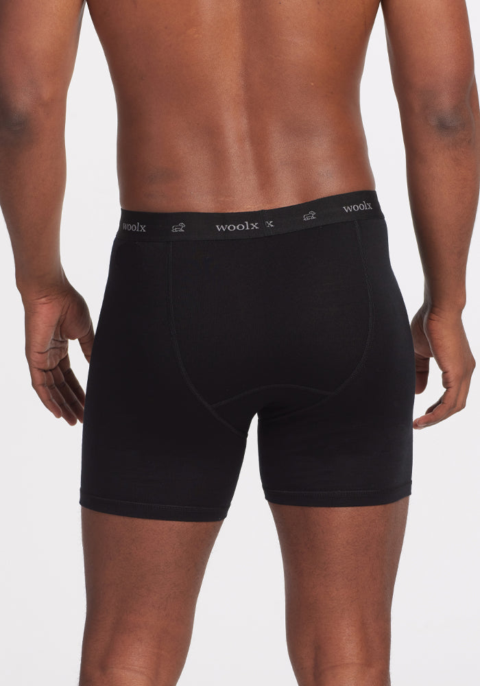 A person is standing with their back to the camera, wearing Woolx Reaction Boxer Briefs in black, featuring "woolx" on the waistband. Crafted from DuraLite Fabric, these briefs provide a perfect blend of comfort and style. The plain white background accentuates their sleek design.