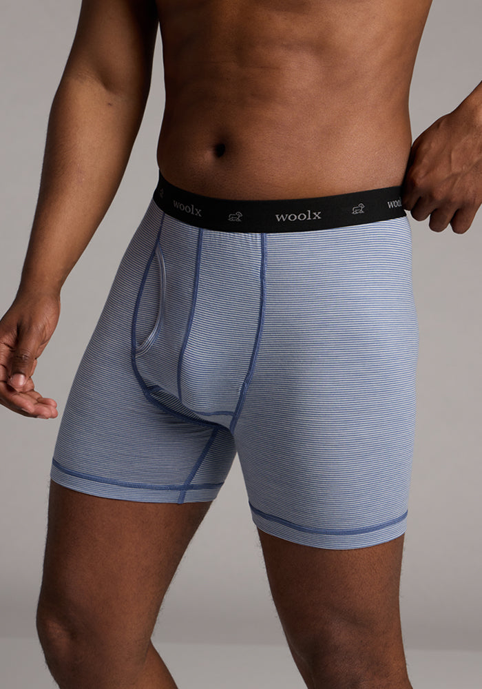 A person models Woolx Reaction Boxer Briefs in Blue Sand Mini Stripe, featuring a light blue striped design and a black waistband with "woolx" in white text, set against a neutral gray background. The torso and legs are visible.