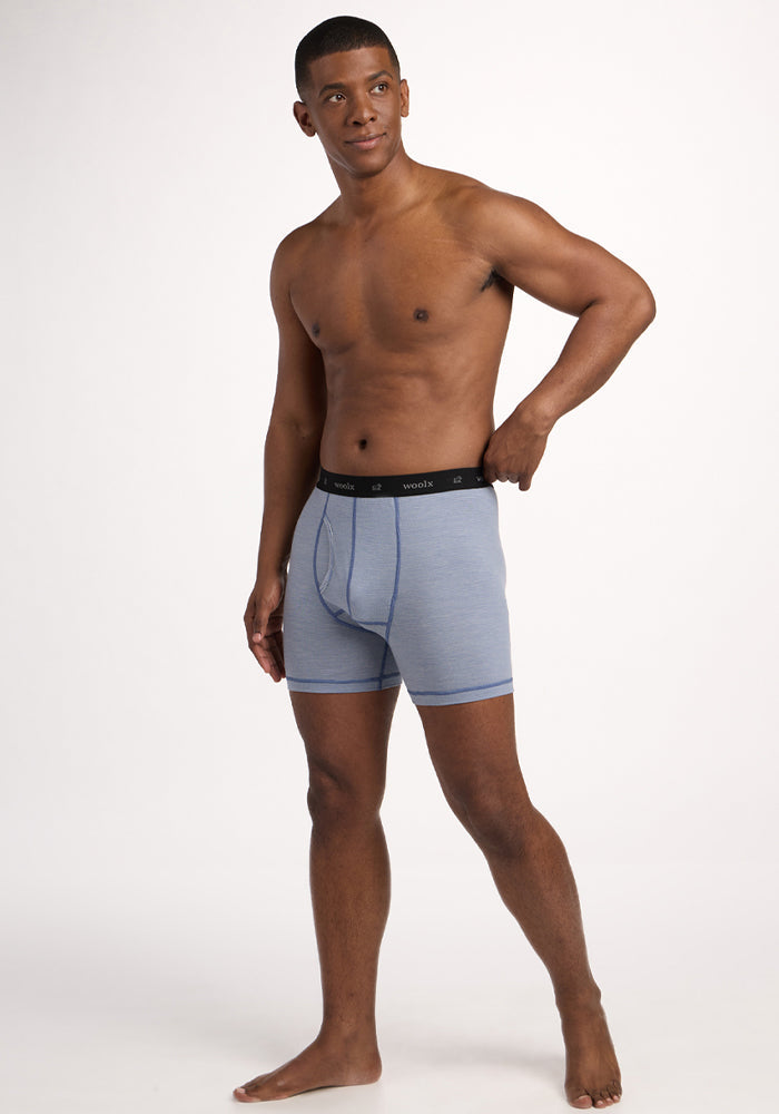 A man stands barefoot against a plain white background, wearing Woolx Reaction Boxer Briefs in Blue Sand Mini Stripe with a black waistband. He looks slightly to the side with one hand on his hip.
