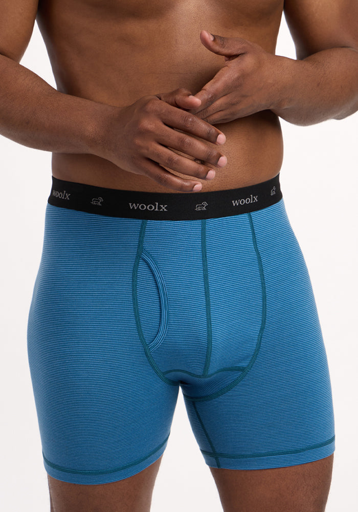 A man stands against a white background wearing Woolx Reaction Boxer Briefs in Coastal Mini Stripe, featuring a blue merino wool fabric and a black waistband with "woolx" in white letters. His face is not visible; only his torso and hands are shown.