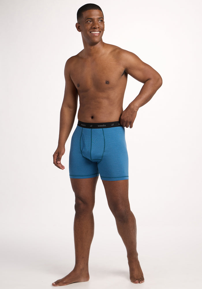 A man smiles in a studio, barefoot and posing against a plain white background, wearing Woolx Reaction Boxer Briefs - Coastal Mini Stripe with a black waistband.