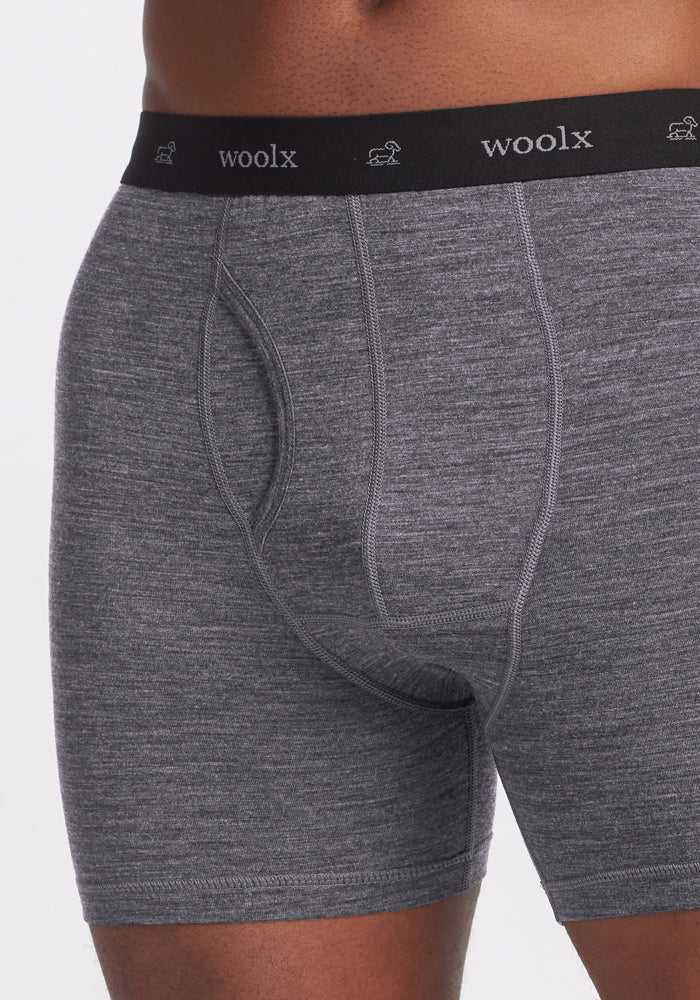 A close-up features someone in Woolx Reaction Boxer Briefs in Graphite Heather, showcasing a black waistband with "woolx" and small sheep icons. Made from Merino Wool, the fabric looks soft and stretchy, offering comfort for everyday wear.