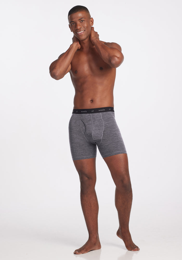 A man stands smiling with hands behind his neck, wearing Woolx Reaction Boxer Briefs in Graphite Heather with a black waistband. Barefoot against a plain white background, he exudes comfort and ease.
