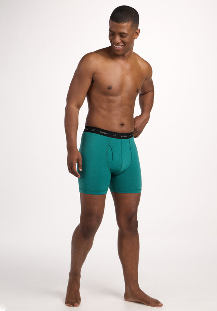 A smiling man stands barefoot against a plain white background, wearing Woolx Reaction Boxer Briefs - Woodland Mini Stripe, featuring a comfortable green merino wool fabric with a black waistband.