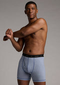 A man wearing Woolx Reaction Boxer Briefs in Blue Sand Mini Stripe, featuring a black waistband, stretches his arm across his chest against a plain, neutral background.