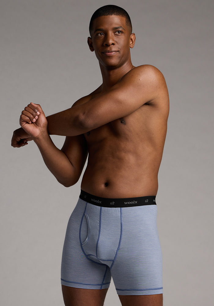 A man wearing Woolx Reaction Boxer Briefs in Blue Sand Mini Stripe, featuring a black waistband, stretches his arm across his chest against a plain, neutral background.