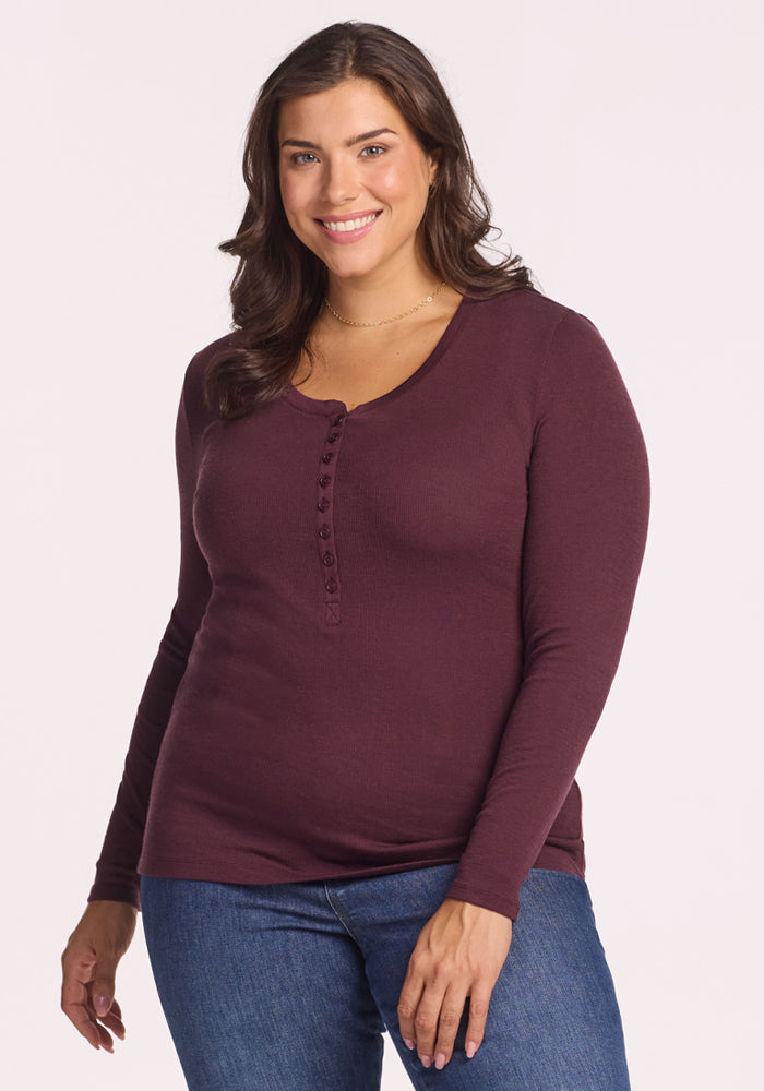 A woman with long brown hair, dressed in the Woolx Reese Ribbed Henley in Chocolate Fig and blue jeans, stands smiling against a plain light background. | Juliana is 5'10", wearing a size XL